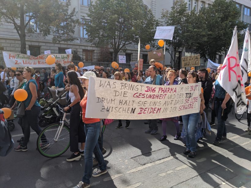 Menschenmasse mit großem Banner in der Hand. Text darauf: "WAS BRINGT BIG PHARMA UM IHR BROT? DIE GESUNDHEIT UND DER TOD! DRUM HÄLT SIE UNS AUF DASS SIE LEBE ZWISCHEN BEIDEM IN DER SCHWEBE."
