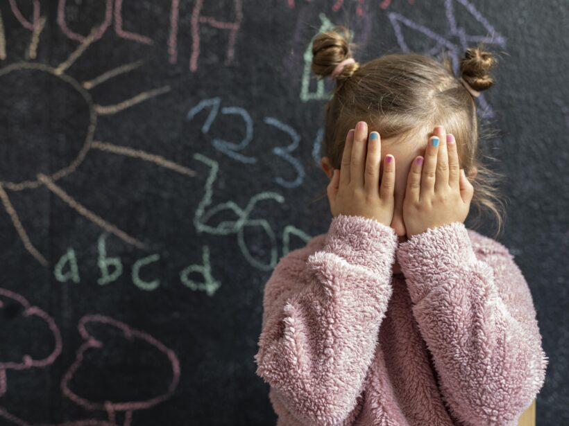 Mädchen, stehend vor einer voll beschrifteten Schultafel, hält sich beide Hände vor ihr Gesicht.